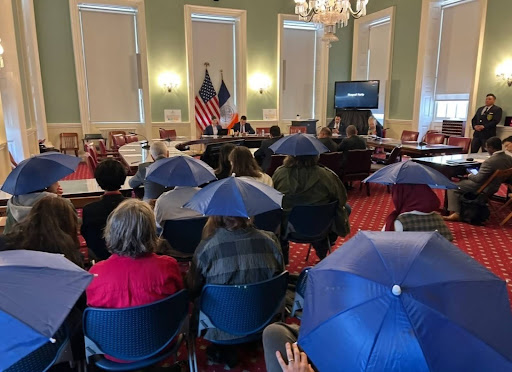 NYC hall hearing room filled with citizens holding umbrellas. 