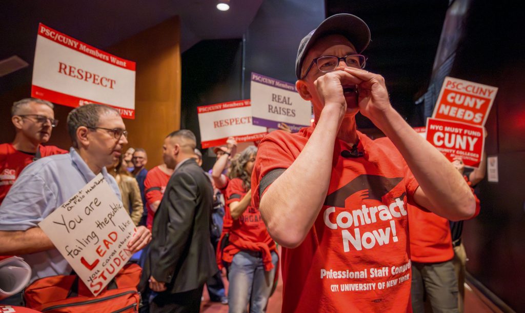 James Davis shouts in a CUNY trustees meeting. Members hold up signs.